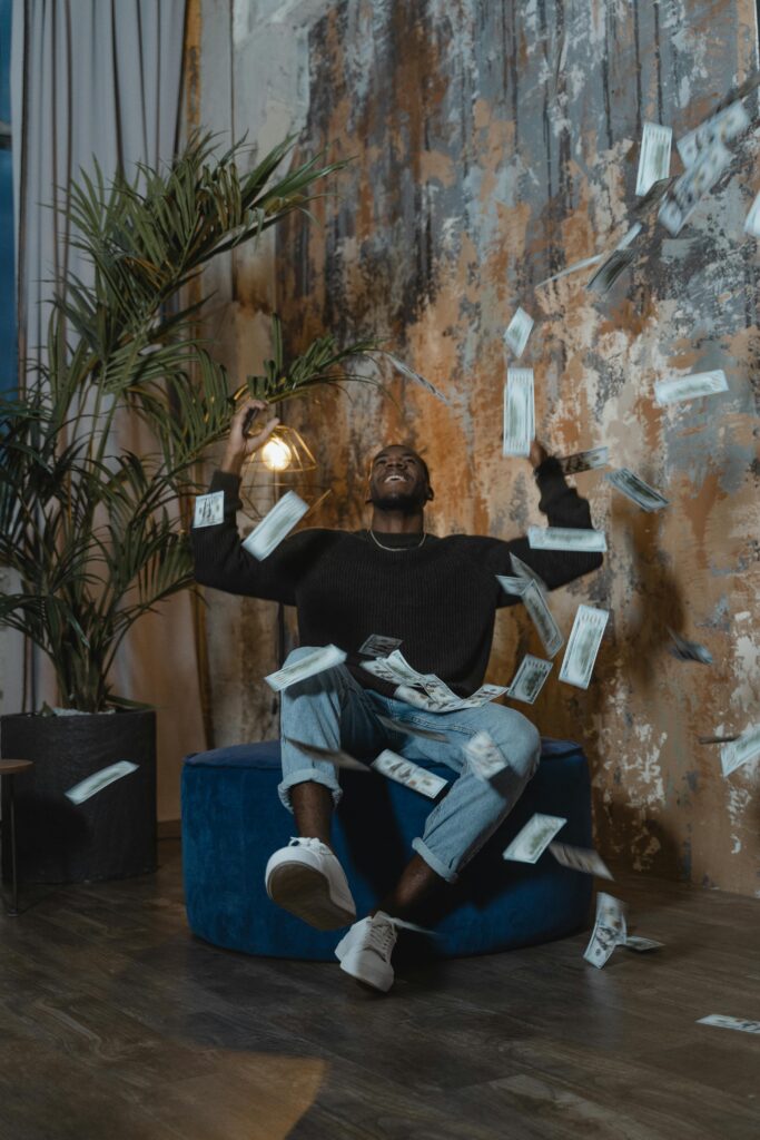Man sitting indoors joyfully celebrating as money falls around him, creating a lively atmosphere.